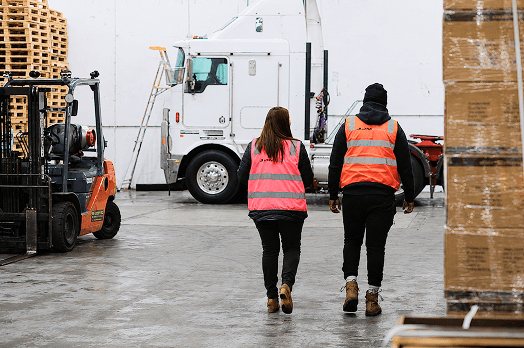 Two warehouse workers in orange safety vests walking toward a white truck in an industrial setting.